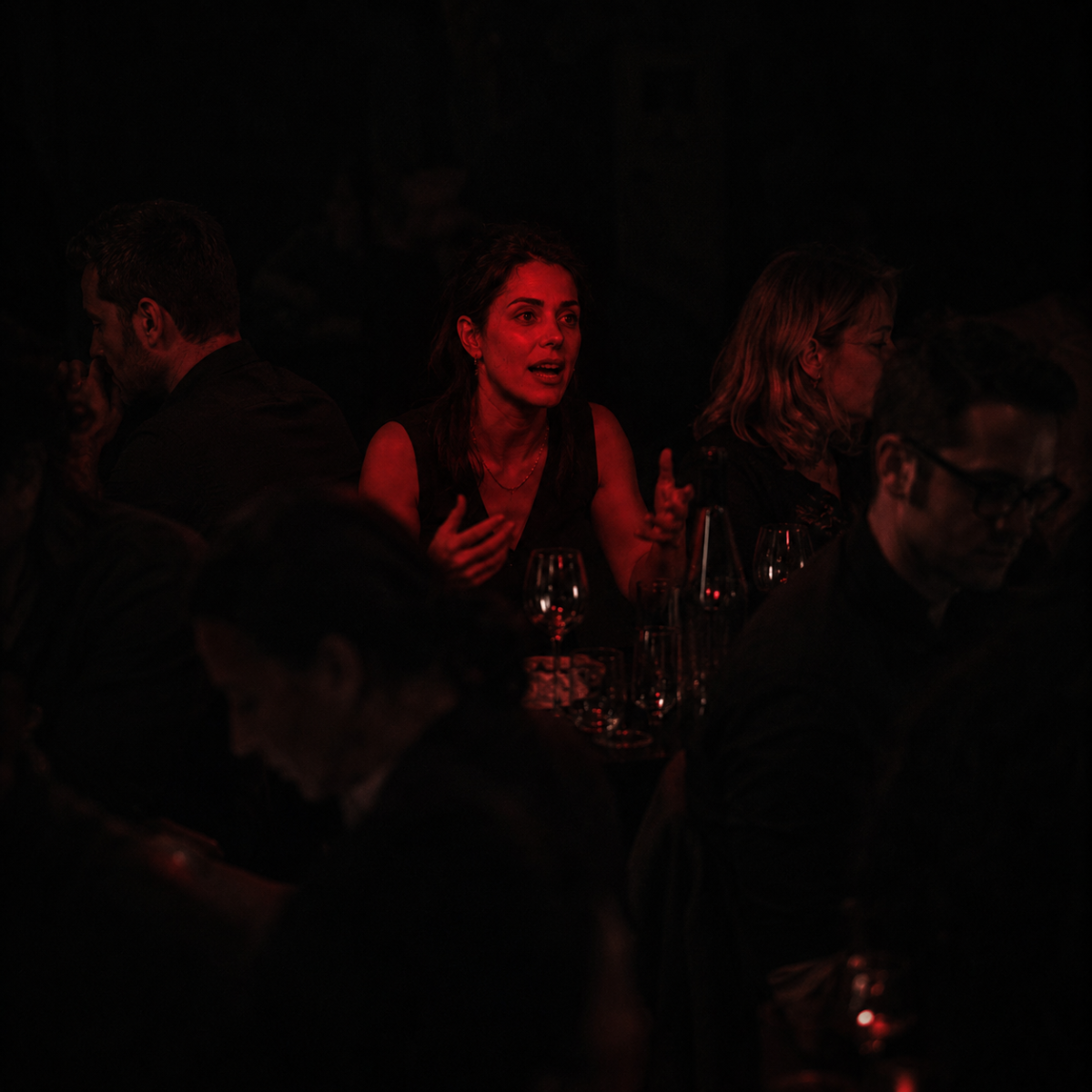 Gaslighting: a woman mid-sentence at a crowded table, the only person lit, while everyone around her looks away