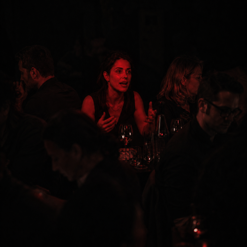 Gaslighting: a woman mid-sentence at a crowded table, the only person lit, while everyone around her looks away